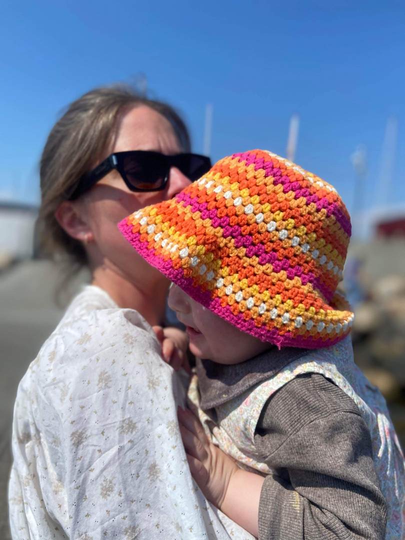 Simone holding her daughter wearing a colorful crocheted sun hat outdoors on a sunny day.