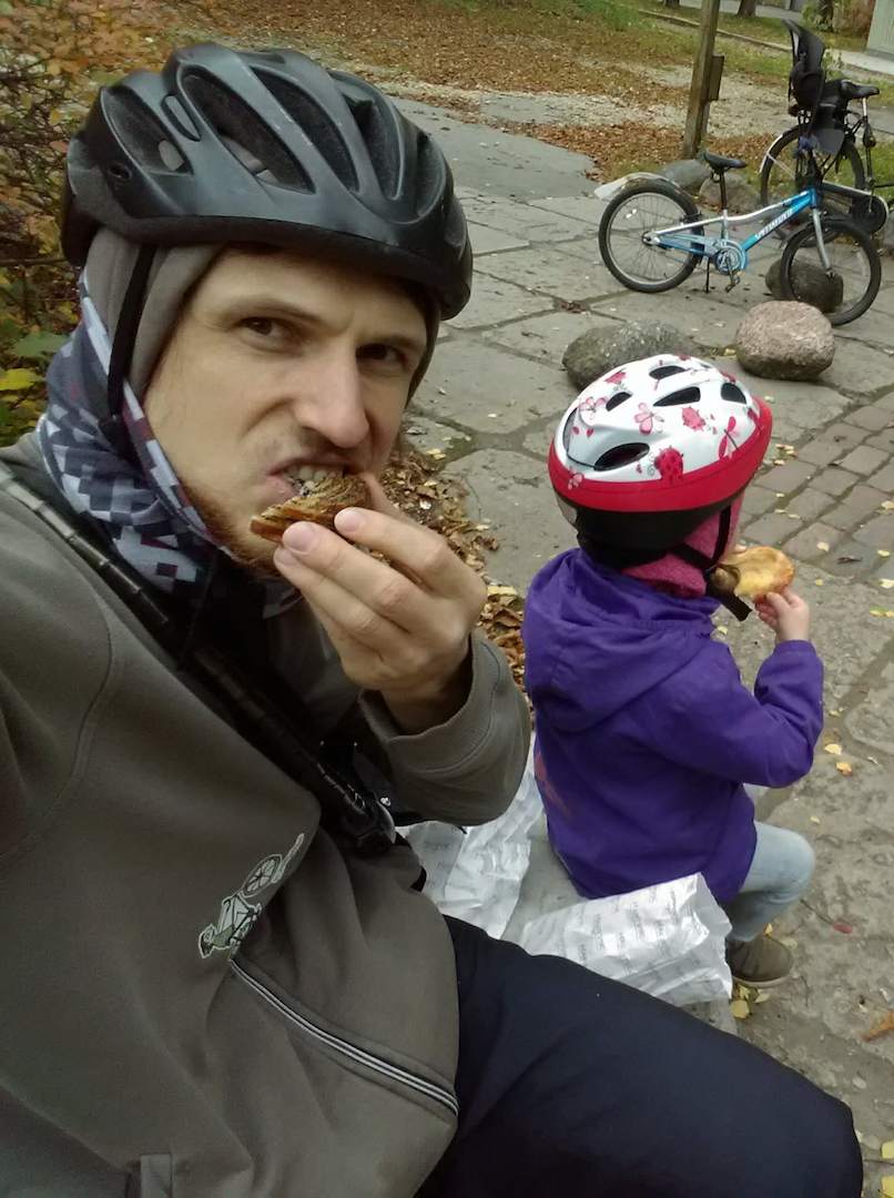 Rainis and his kid wearing bicycle helmets while enjoying a snack during a bike ride.