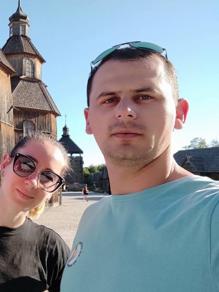 Alexey and his wife posing for a selfie in front of a historic wooden church.