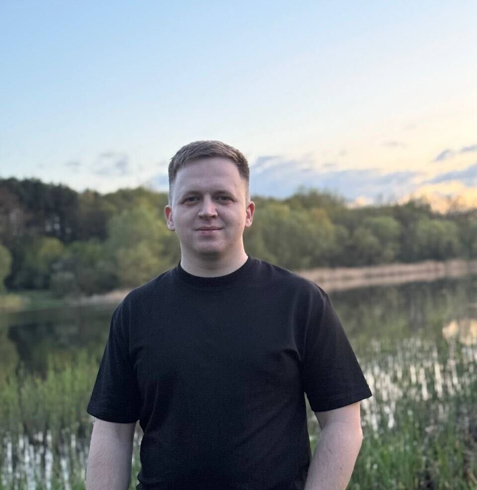 Maksym standing by a lakeside at sunset wearing a black T-shirt.